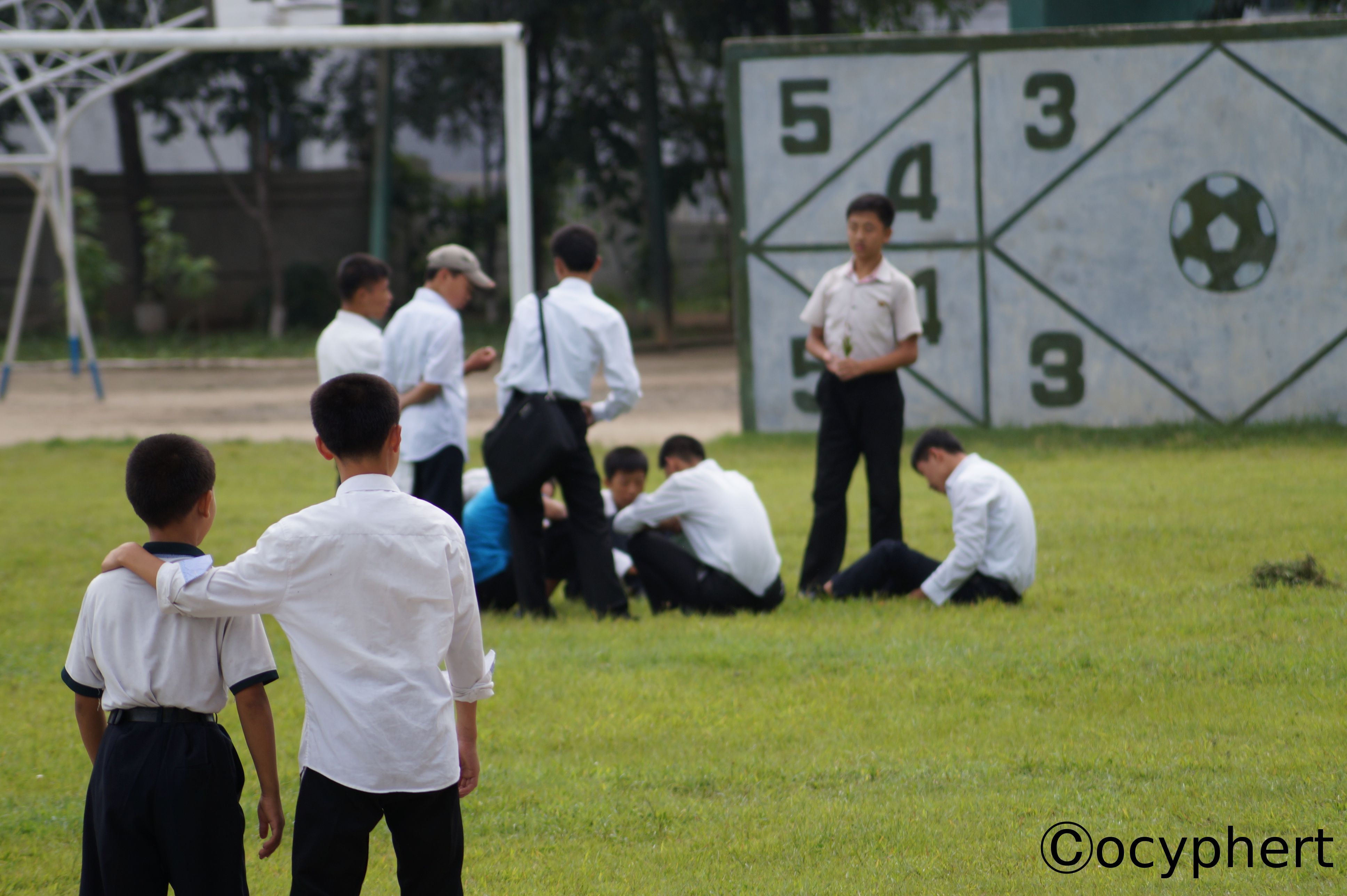 Eine Gruppe Kinder sitzt auf dem Rasen des Pausenhofes. Im Hintergrund sieht man ein Fußballtor, im Vordergrund einen Jungen, der einem etwas kleineren Jungen die Hand auf die Schulter legt.