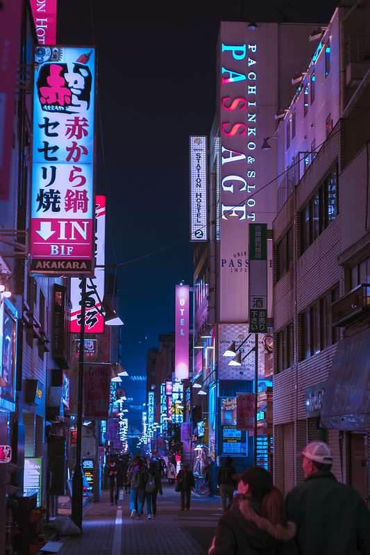 Shinjuku in Tokio bei Nacht. Die Häuser haben an der Außenwand viele neon-licht-Schilder, welche in die Straße reichen.