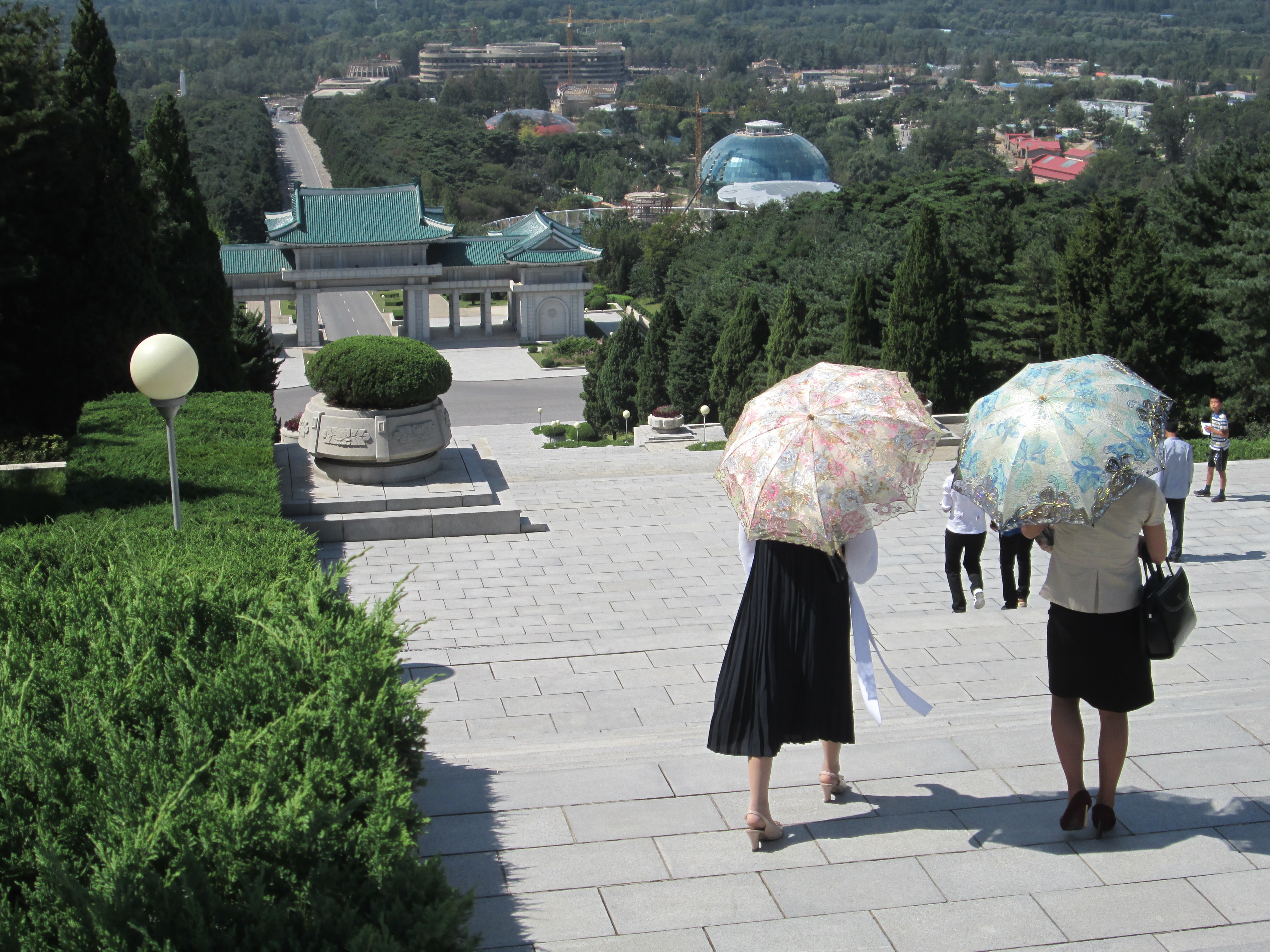 Blick über Treppenstufen und eine Aussicht über Pjöngjang. Im Vordergrund sieht man zwei Frauen von hinten, welche jeweils einen Sonnenschirm tragen.