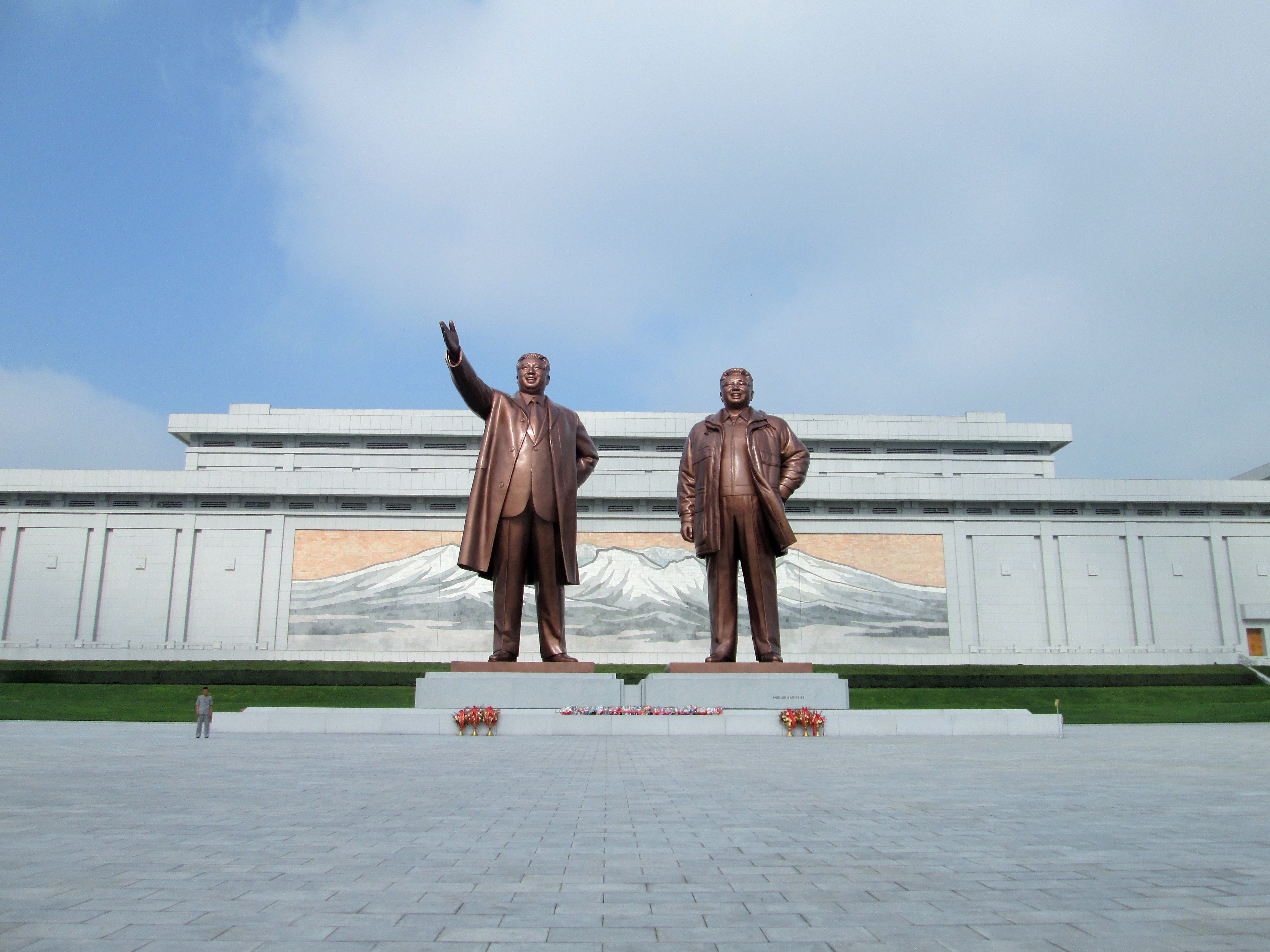 Die monumentalen Bronze-Statuen von Kim Jong Il und Kom Jong Un erstrecken sich auf einem riesigen bepflasterten Platz in die Höhe. Vor den Statuen wurden Blumen abgelegt. Ein Mensch neben den Stauen zeigt dass die Statuen selbst mindestens 7 Menschen hoch sein muss.