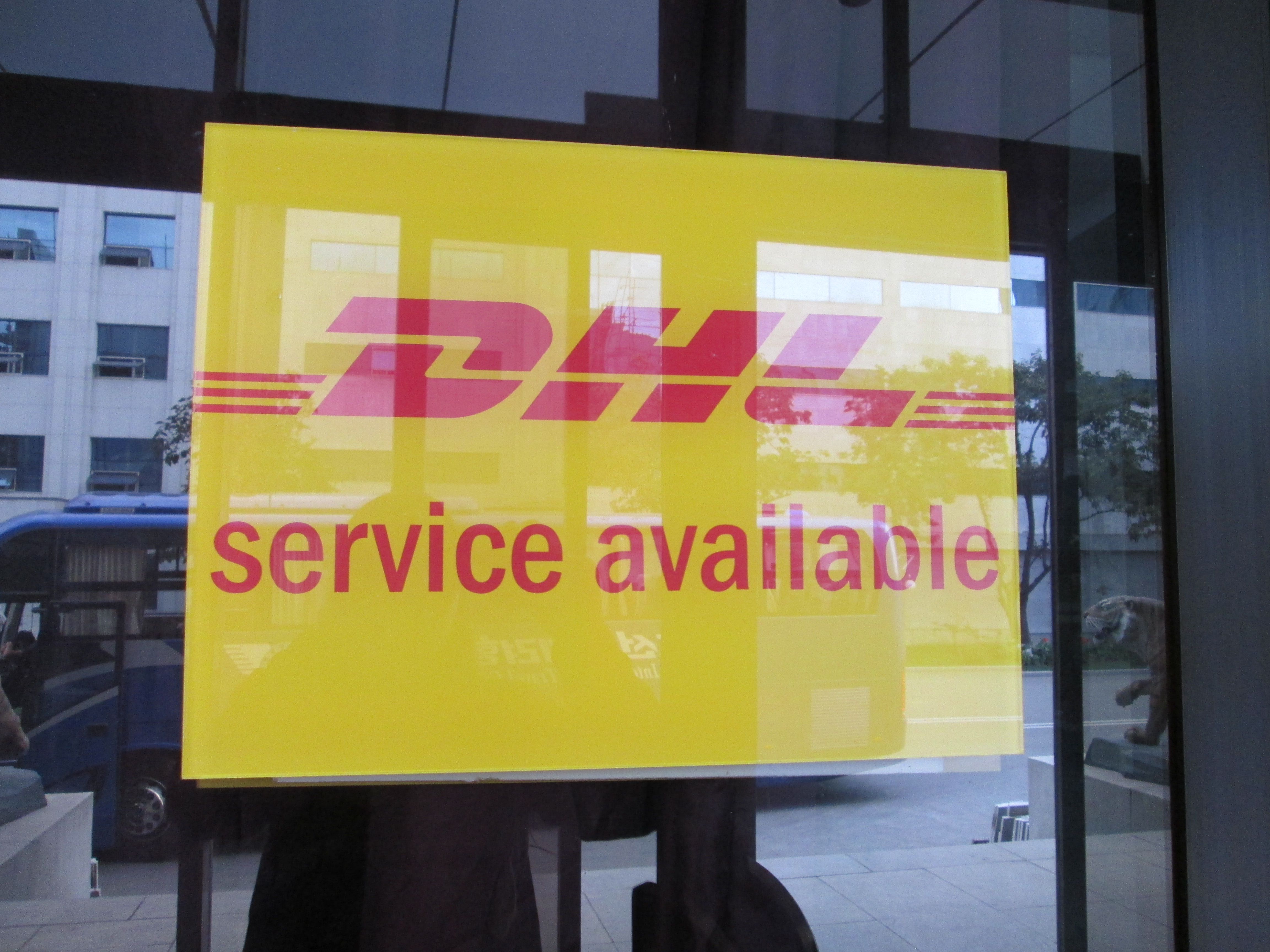 Hinter einer Glasscheibe hängt ein Schild mit der Aufschrift: "DHL verfügbar" mit DHL-Logo
